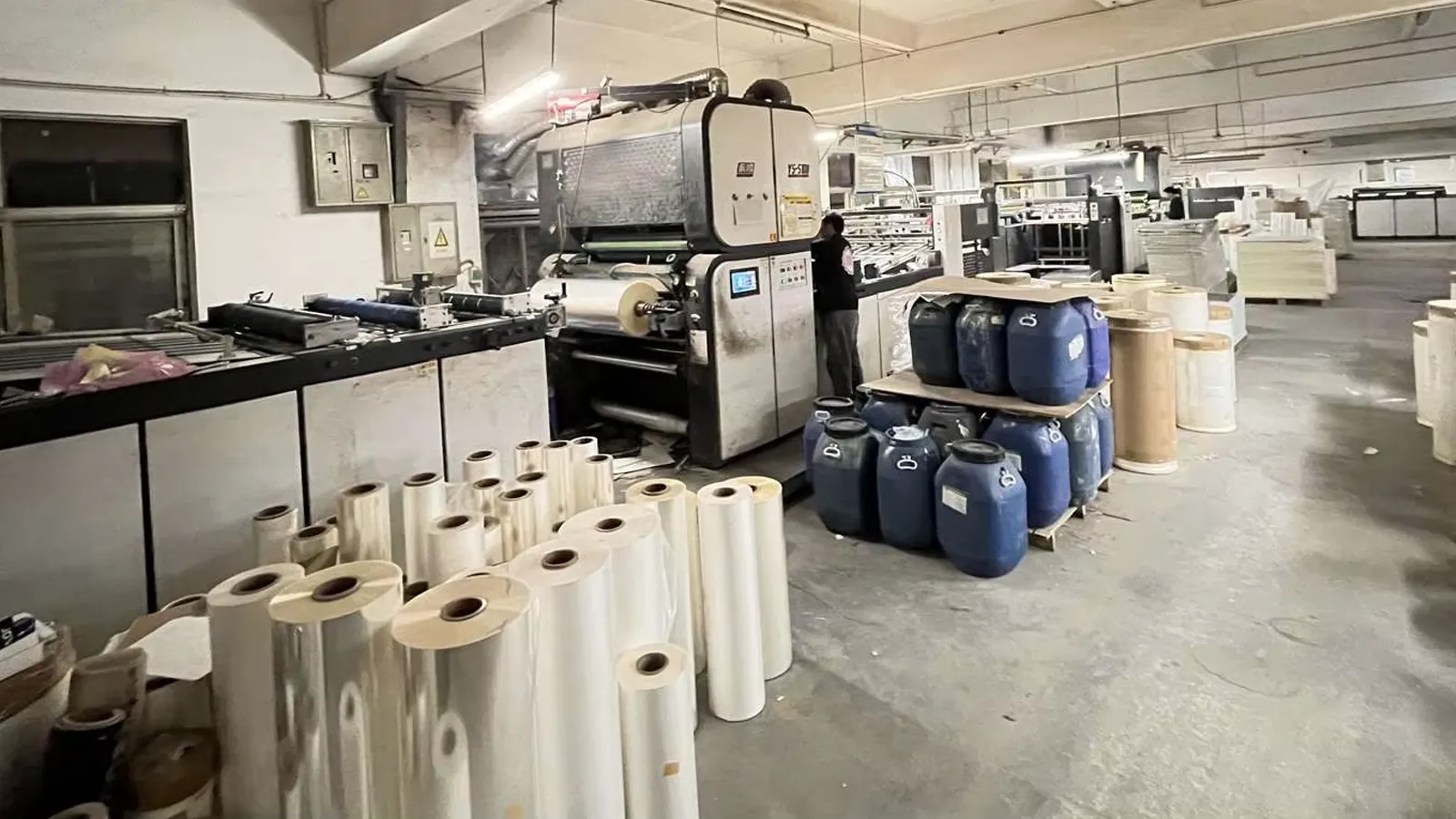 Lamination machine in a paper packaging factory with rolls of film and production materials on the workshop floor.